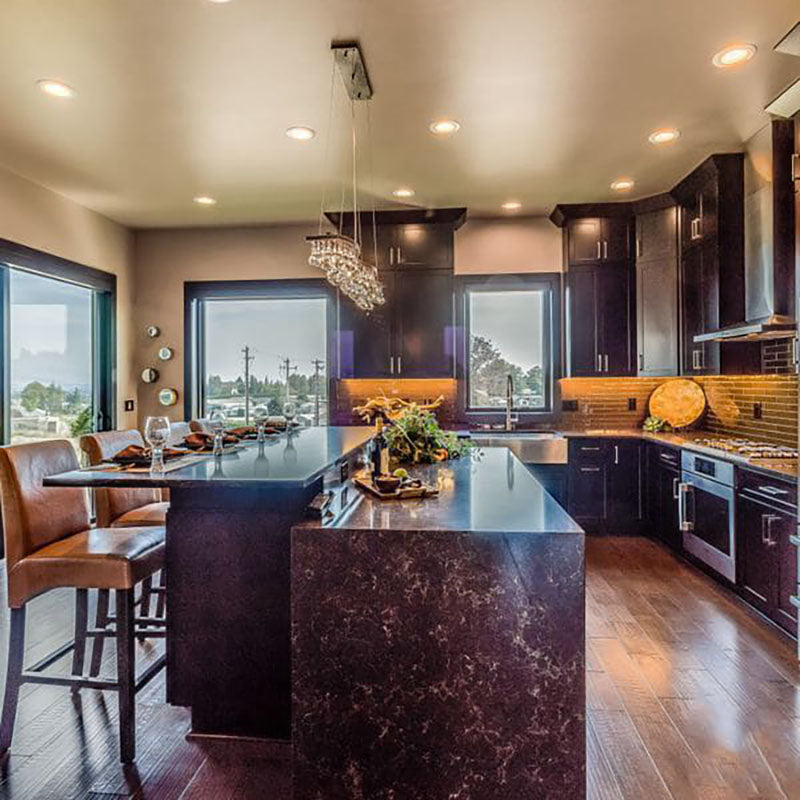 View from the side of a granite kitchen island with light brown leather chairs, with a large lighting fixture hanging above and dark kitchen cabinets, designed by Kate Loeb, Designer at Standard Paint & Flooring.