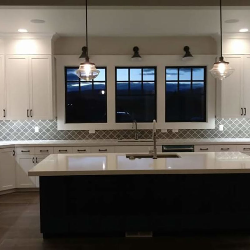 A renovated kitchen designed by Standard Paint & Flooring, with white cabinets and a grey and white accent back splash tile, with a kitchen island with white marble countertop and dark wood siding. 
