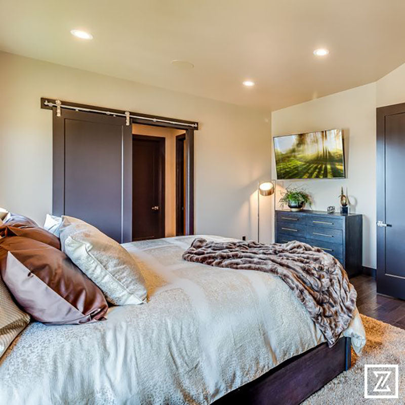 A bedroom with hardwood flooring, a plush area rug under a king sized bed with beige and gold blankets and pillows, with dark wood barn style doors on the closet, designed by Kate Loeb, Designer at Standard Paint & Flooring. 