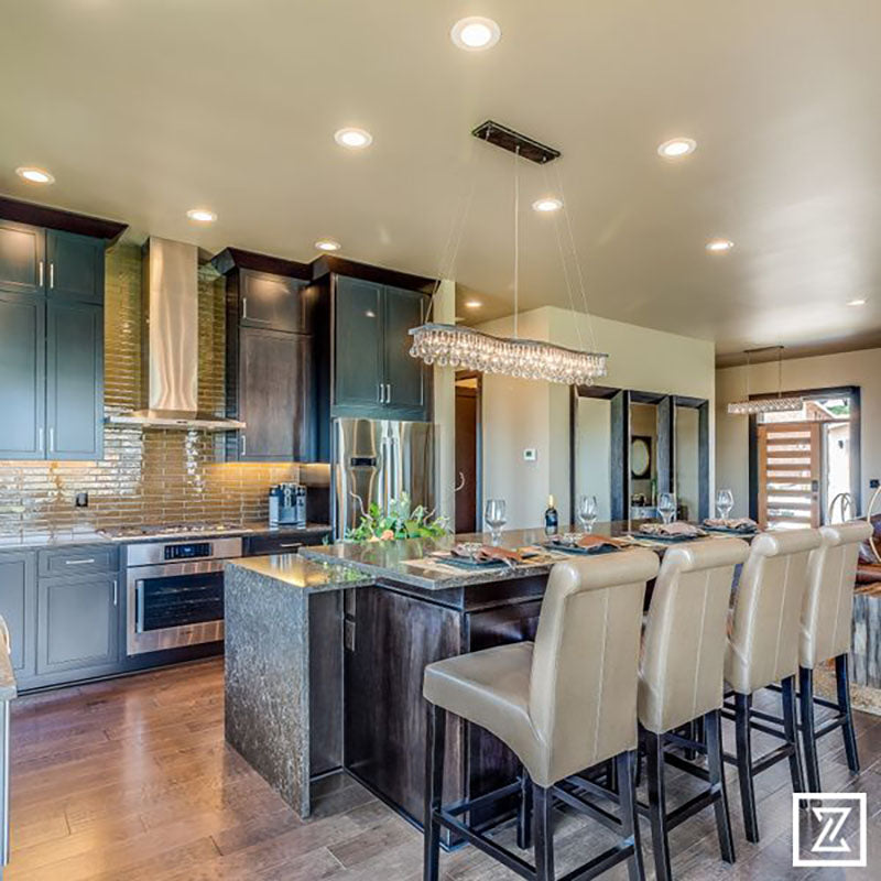 View from behind a kitchen island with a granite countertop, dark wood siding and light brown leather chairs, with a large lighting fixture hanging above and dark kitchen cabinets, designed by Kate Loeb, Designer at Standard Paint & Flooring. 