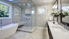 Bathroom with a glass panel shower, white bath tub, white marble countertop with a white vase and white flowers, and light beige and white marbled tile flooring. 