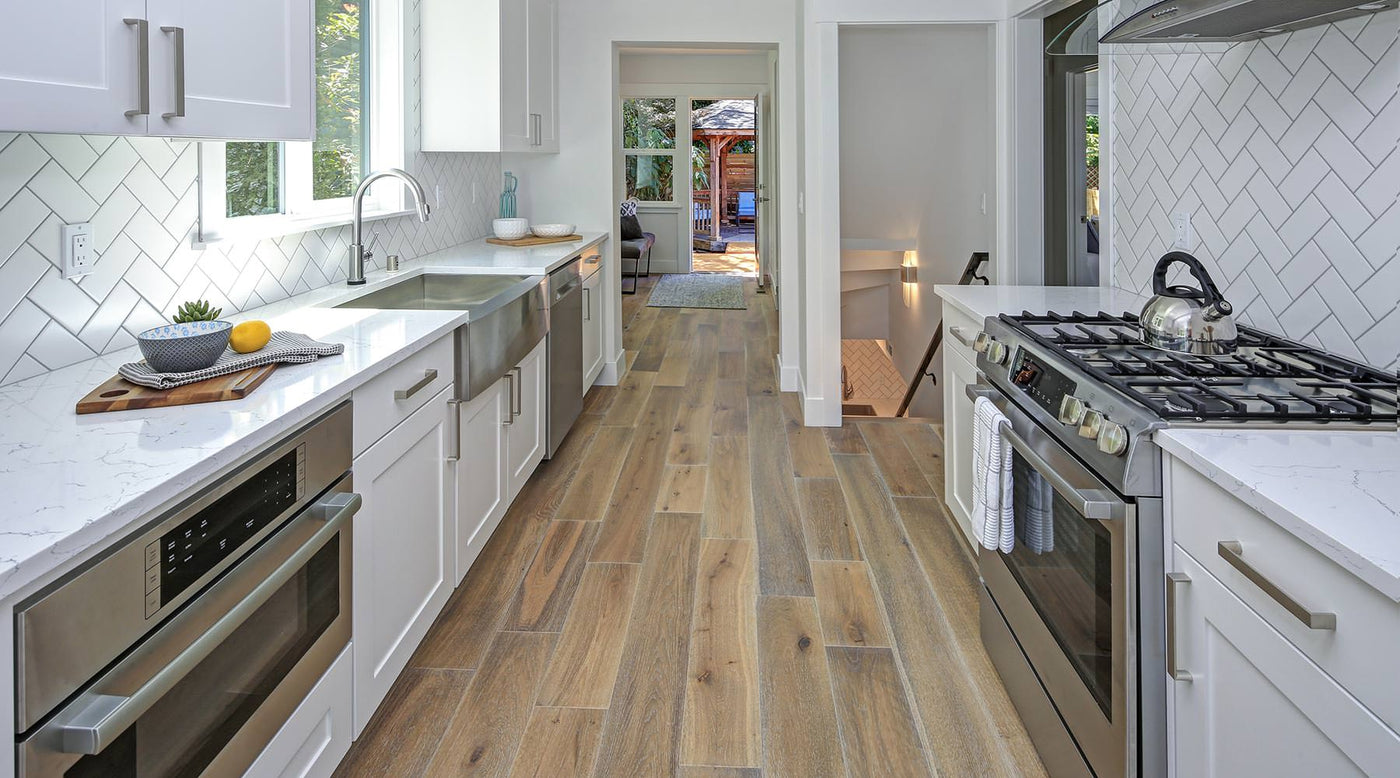 A galley kitchen with white countertops and cabinets and stainless steel appliances on each side, with an alleyway of vinyl plank flooring.