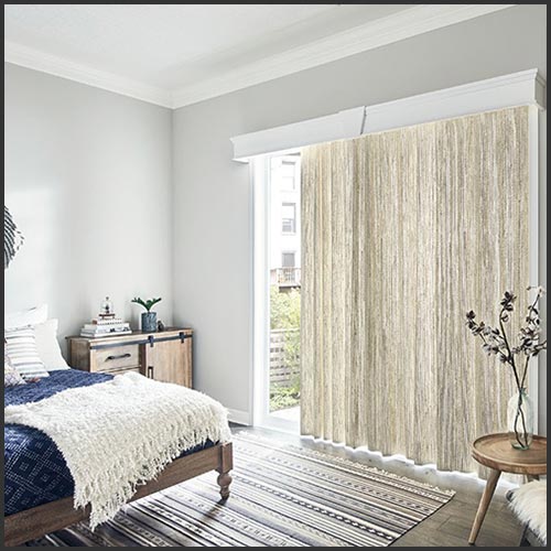 A bedroom with long cream colored window curtains, a patterned carpet and a bed with a dark blue comforter and a cream blanket.